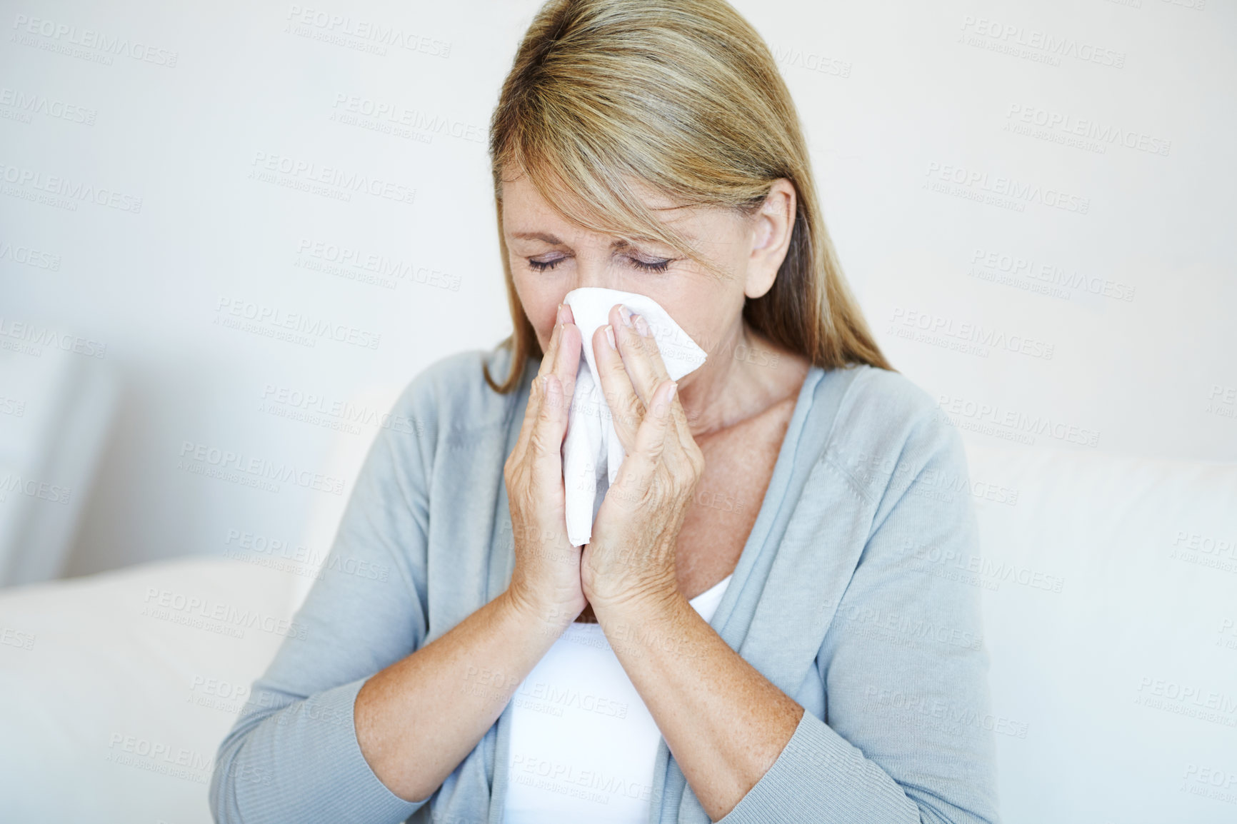 Buy stock photo A mature woman sitting in her livingroom blowing her nose in a tissue