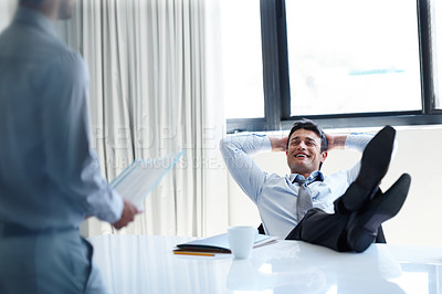 Buy stock photo A laidback businessman with his feet up on his desk as a coworker stands by