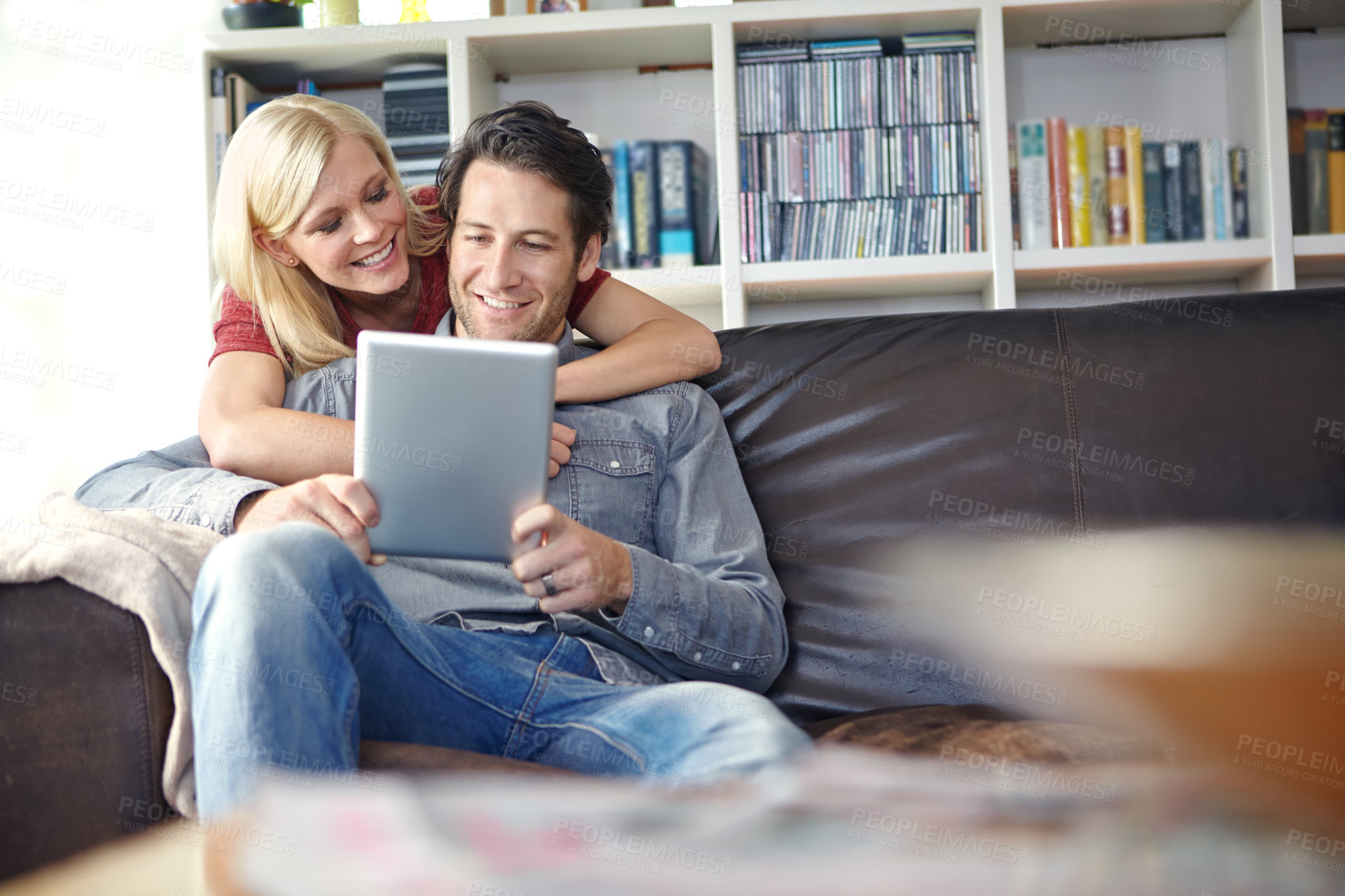 Buy stock photo Shot of an attractive young woman embracing her boyfriend while he uses a digital tablet