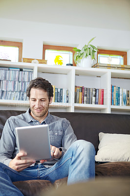 Buy stock photo Shot of a handsome young man sitting on a couch while using a digital tablet