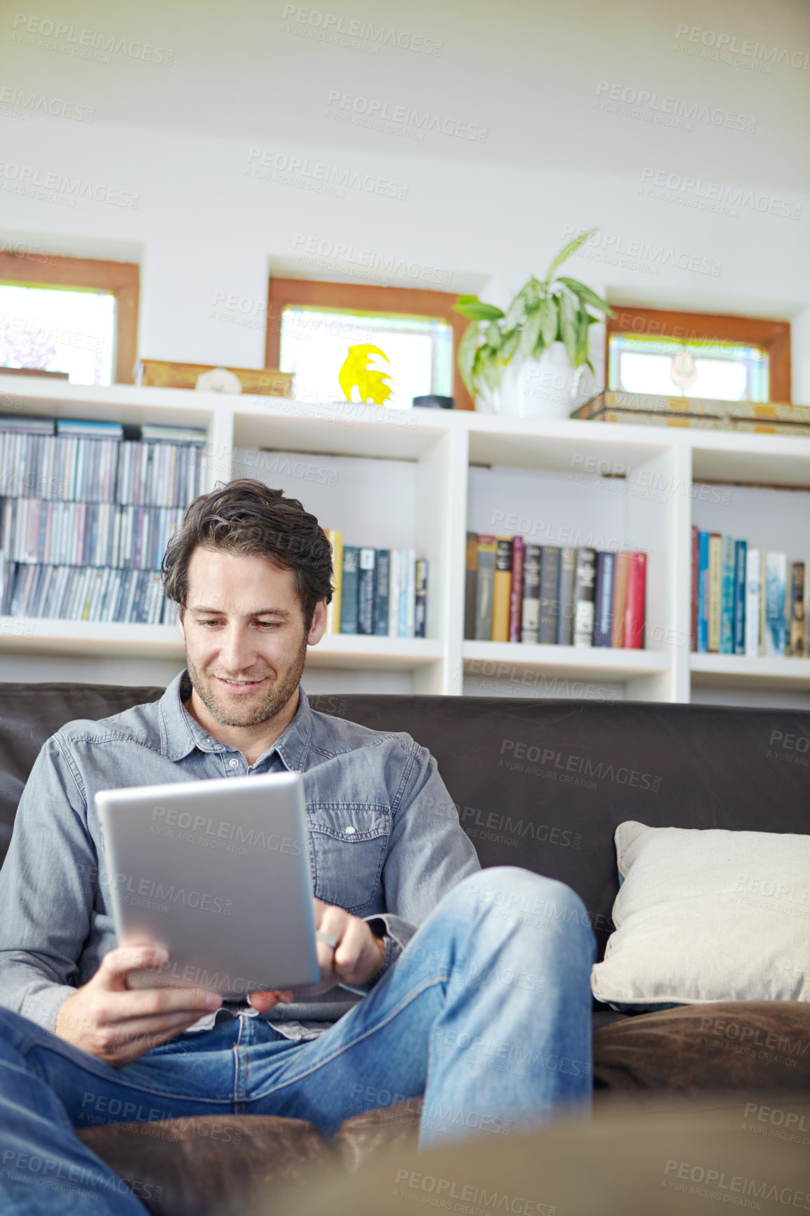Buy stock photo Shot of a handsome young man sitting on a couch while using a digital tablet