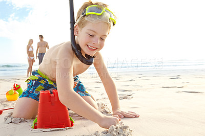 Buy stock photo Shot of a young boy building a sandcastle on the beach while his parents stand in the background