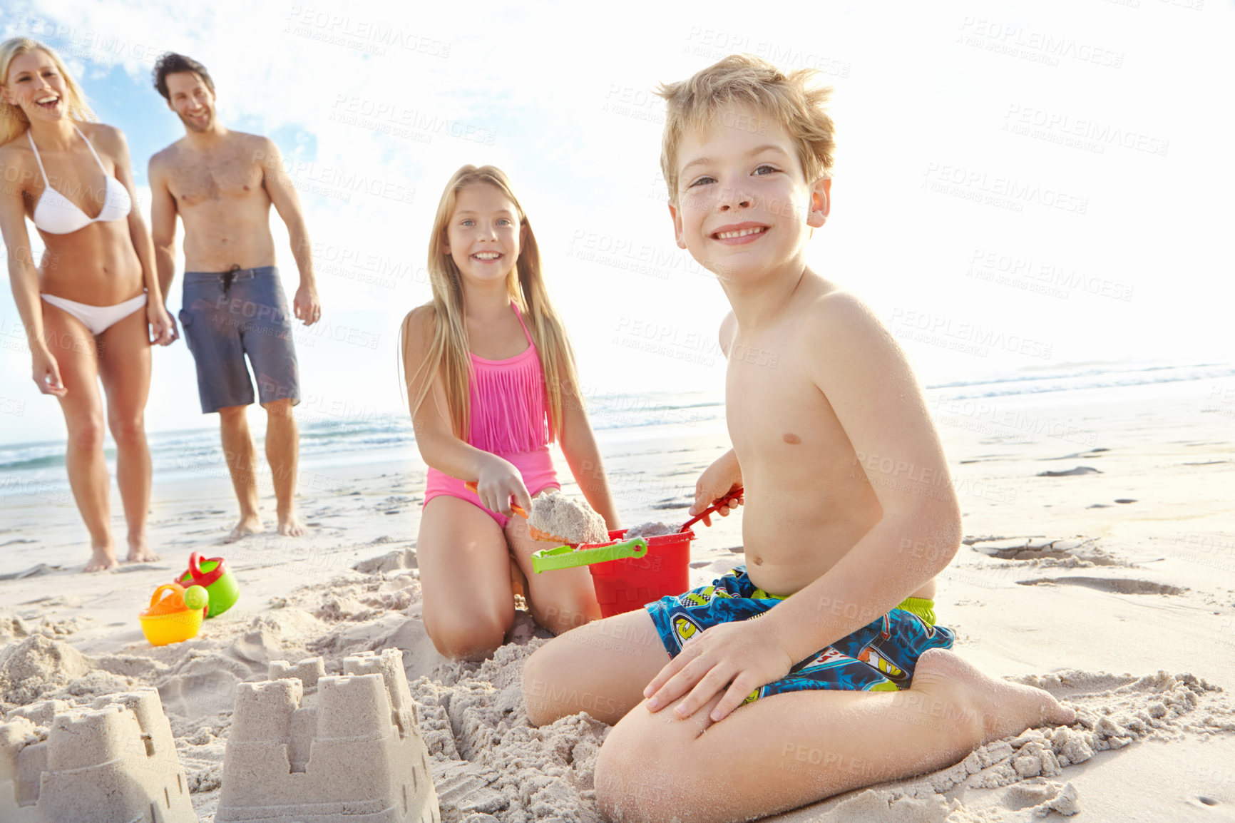 Buy stock photo Shot of a young brother and sister building a sandcastle on the beach while their parents stand in the background