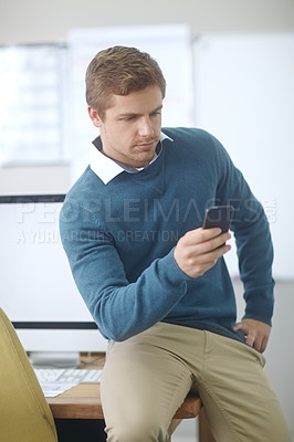 Buy stock photo Shot of a handsome young man using his cellphone while sitting on his desk in an office