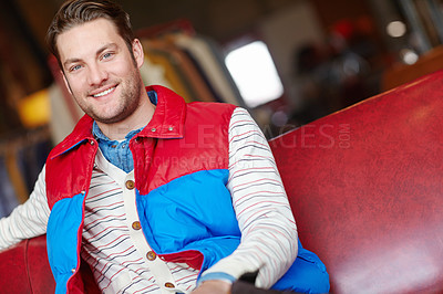 Buy stock photo Portrait of trendy man in bright thrifted jacket sitting inside a fashion store. Young attractive male wearing casual stylish vintage puffer and a striped retro cardigan. Stylish model showing trends
