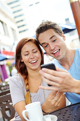 Buy stock photo A happy couple laughing at something on a cellphone while having coffee at a cafe