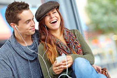 Buy stock photo A pretty girl sharing her earphones with her boyfriend as they listen to music from her phone
