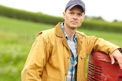 Buy stock photo Portrait of a serious farmer with his arm resting on the hood of his tractor while he stands in an open field