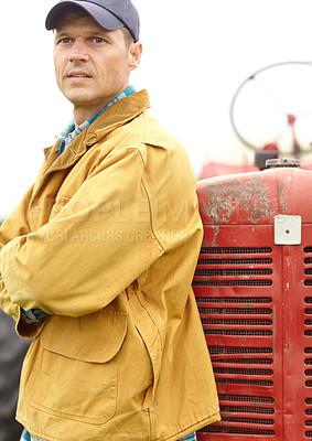 Buy stock photo Shot of a farmer standing next to his tractor with his arms crossed