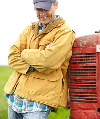 Buy stock photo Shot of a smiling farmer standing next to his tractor with his arms crossed and looking down