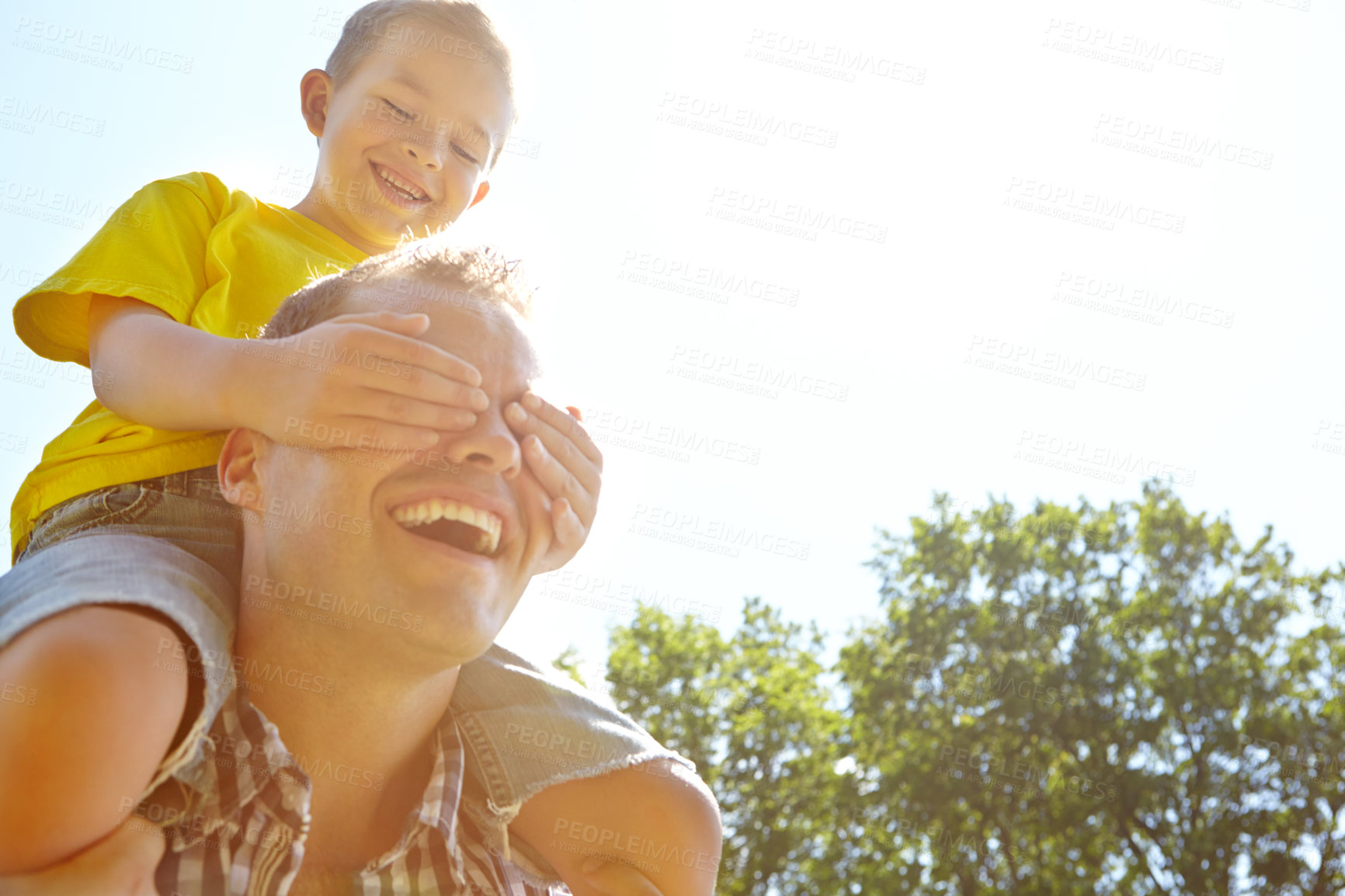 Buy stock photo Low angle shot of a young father with his son sitting on his shoulders and covering his eyes