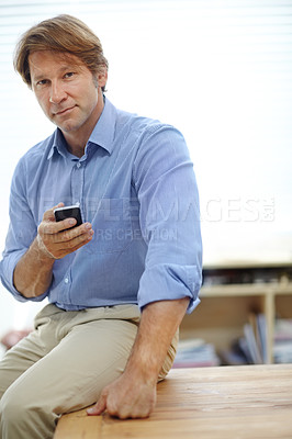 Buy stock photo Portrait of a mature businessman sending a text while leaning on a table in his office