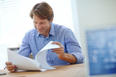 Buy stock photo Shot of a smiling businessman looking over some paperwork while sitting at his desk