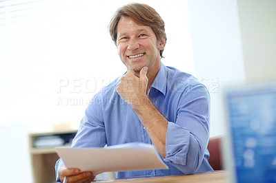 Buy stock photo Shot of a smiling businessman looking over some paperwork while sitting at his desk