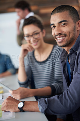 Buy stock photo Portrait of two coworkers sitting at a table in an office with colleagues in the background
