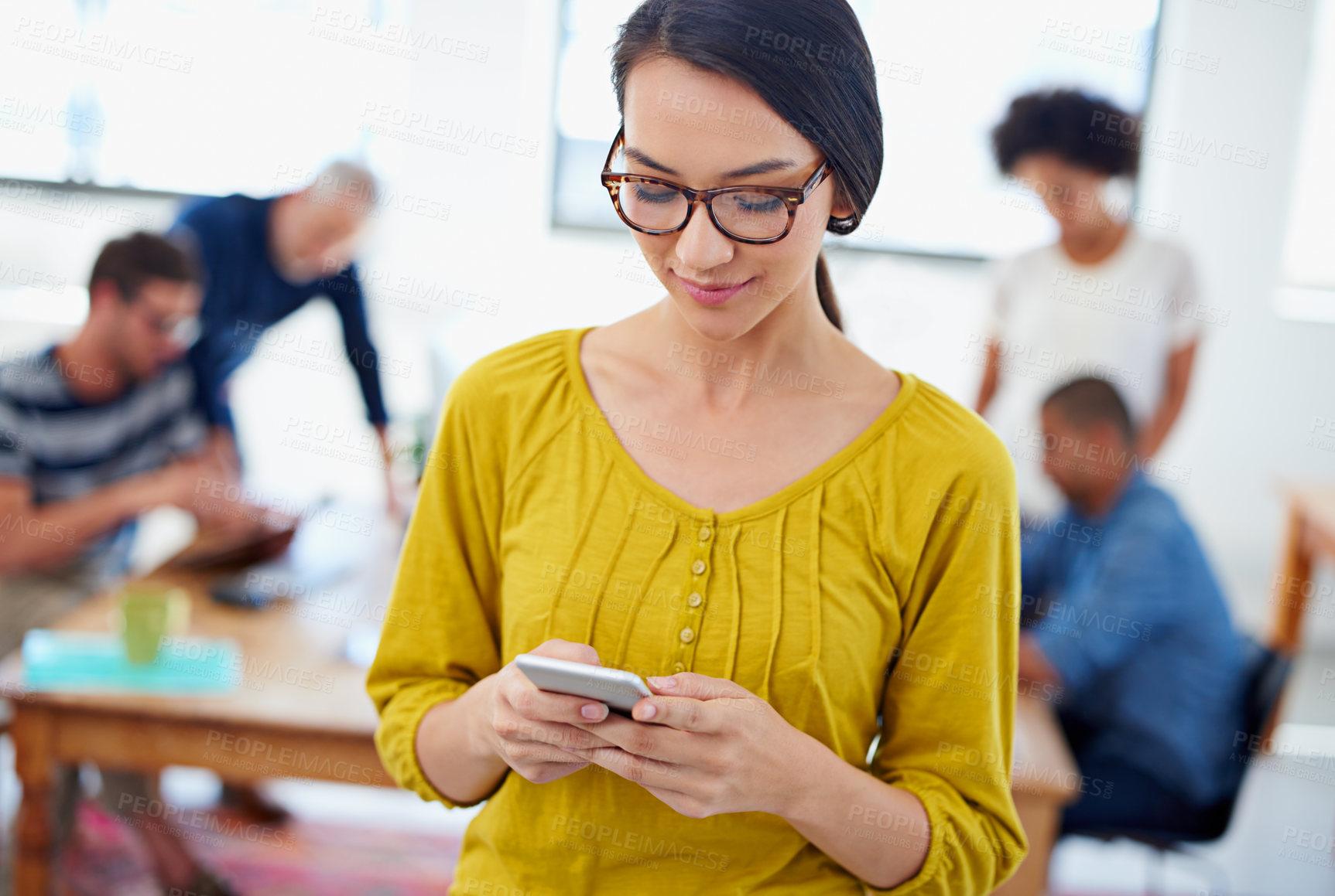 Buy stock photo Young woman texting on her phone with colleagues working behind her