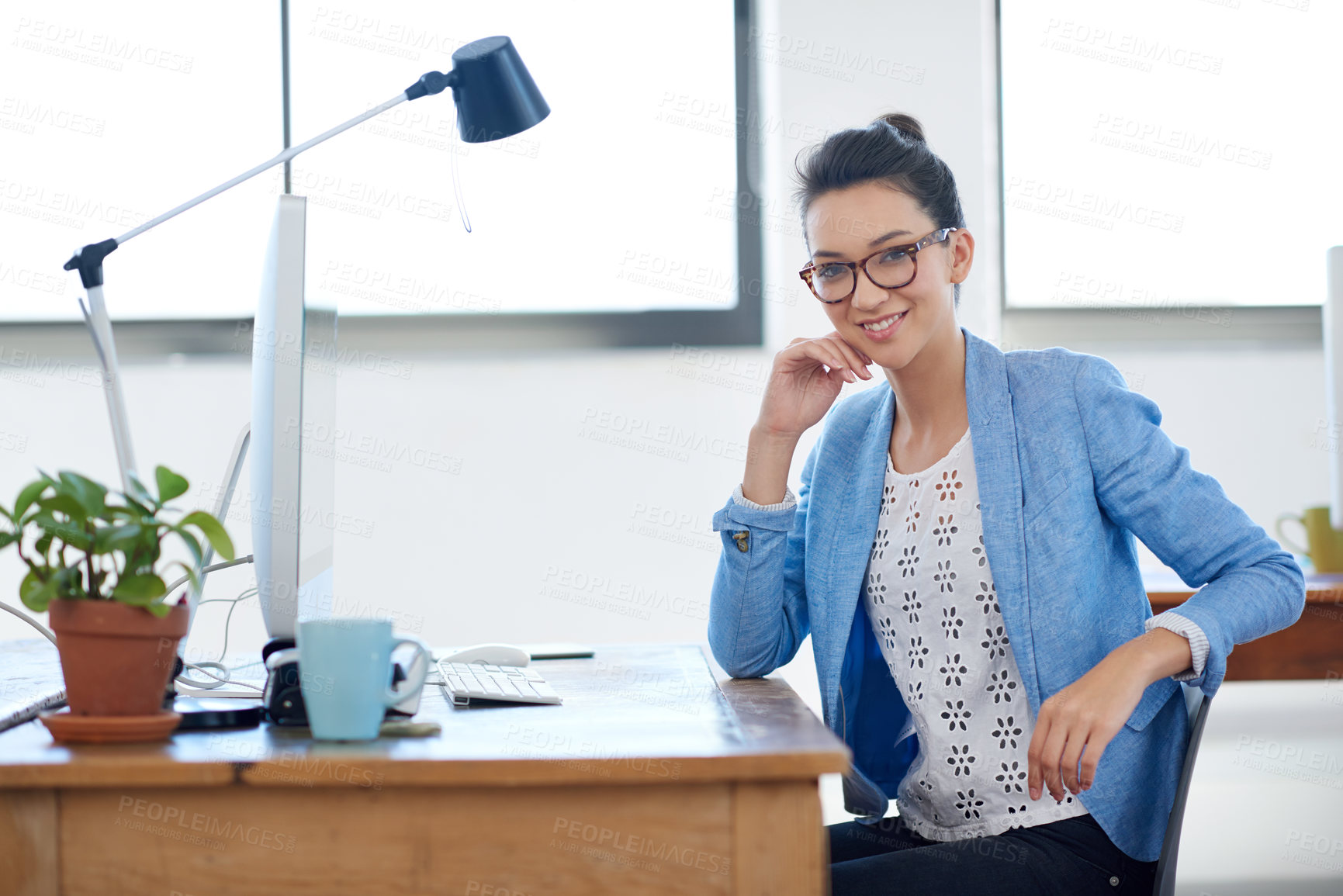Buy stock photo Smiling young woman sitting at her desk in a bright office space