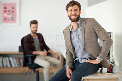 Buy stock photo Handsome young designer smiling at the camera while sitting on the edge of his desk