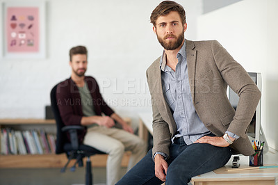 Buy stock photo Serious young businessman sitting on the edge of his desk with a colleague in the background
