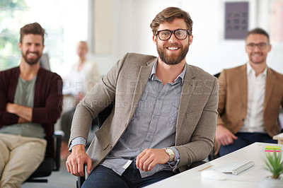 Buy stock photo Smiling young professional sitting at his desk with colleagues in the background
