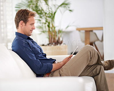 Buy stock photo Shot of a handsome man using his tablet while sitting on the sofa at home