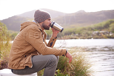 Buy stock photo Shot of a handsome man sitting by a lake drinking from a thermos