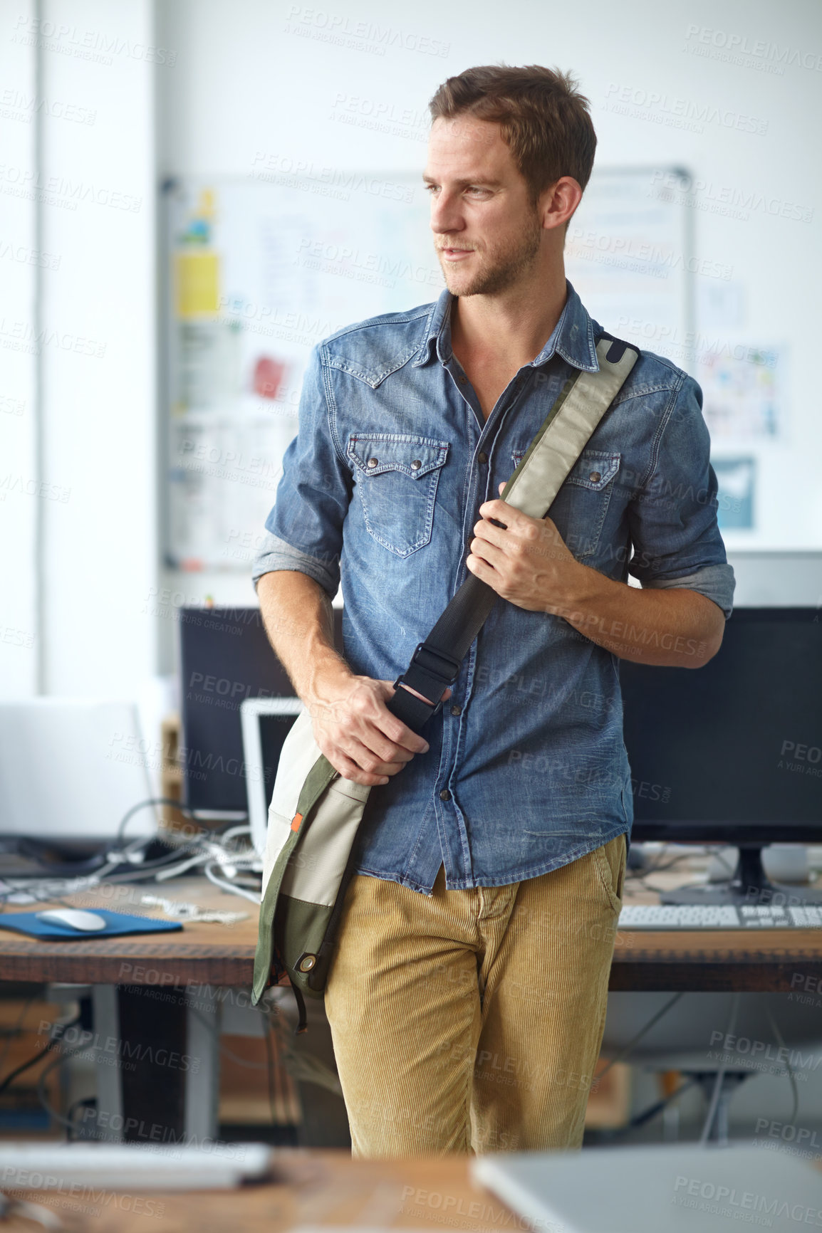 Buy stock photo A handsome young man with a satchel leaning against a table in his office