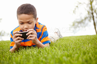 Buy stock photo A little boy engrossed on playing a game on his smartphone while lying outdoors