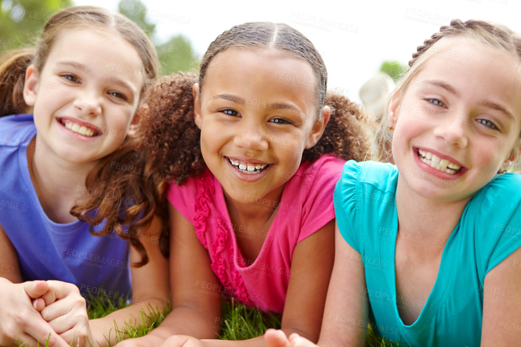 Buy stock photo Three multi-ethnic young girls lying on the grass in a park smiling at the camera