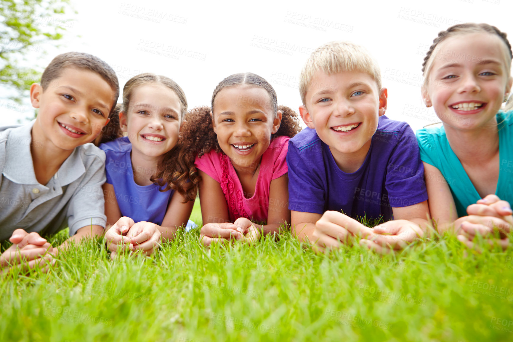 Buy stock photo A multi-ethnic group of children lying on the grass in a park smiling at the camera