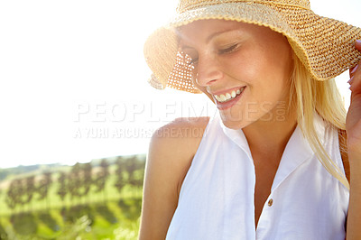 Buy stock photo A lovely young woman wearing a sunhat smiling and looking down as she stands in the countryside