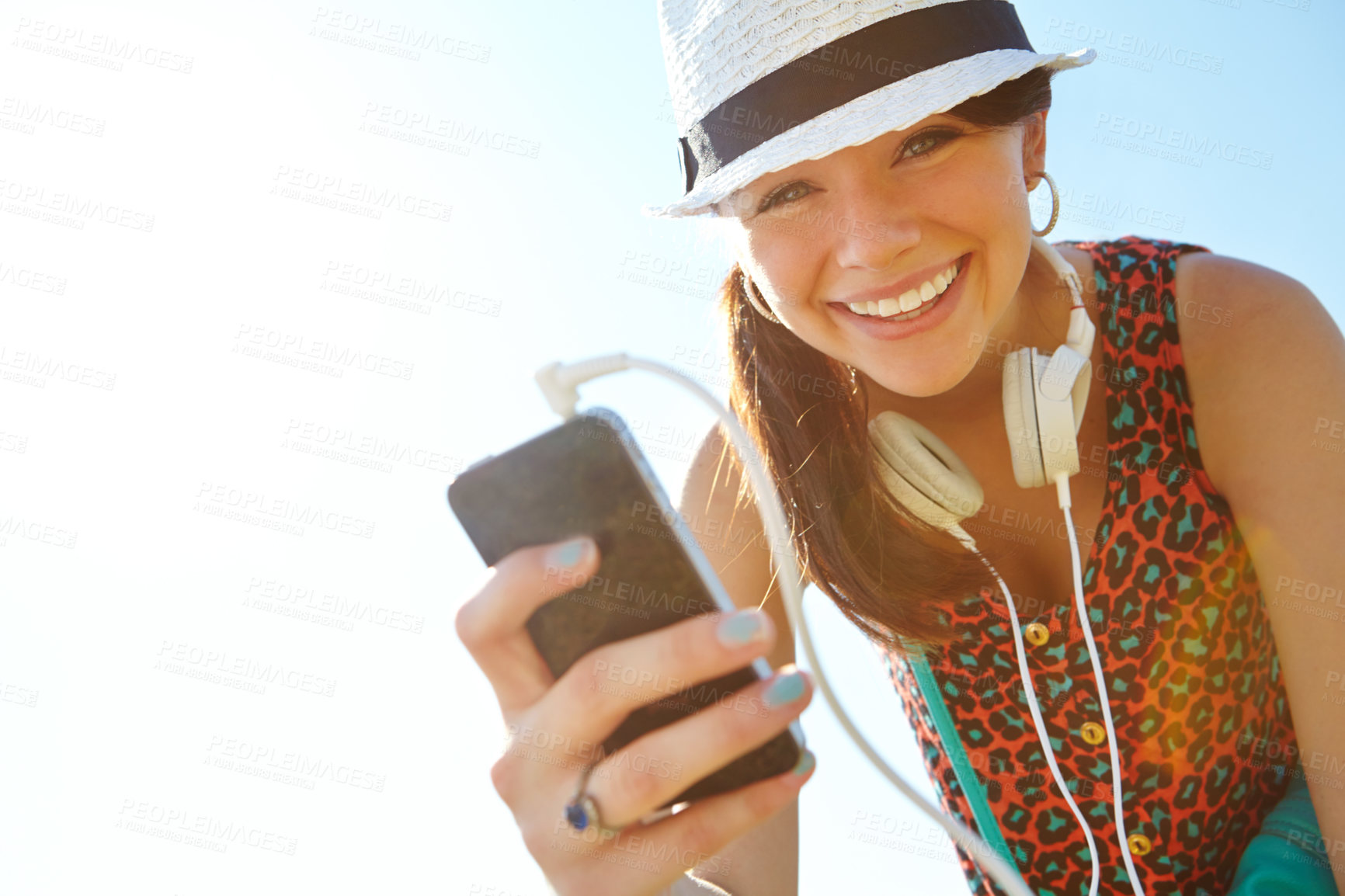 Buy stock photo A happy teenage girl smiling while holding a smartphone with her headphones attached to it