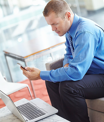 Buy stock photo A young caucasian businessman using his smartphone with his laptop before him as he works