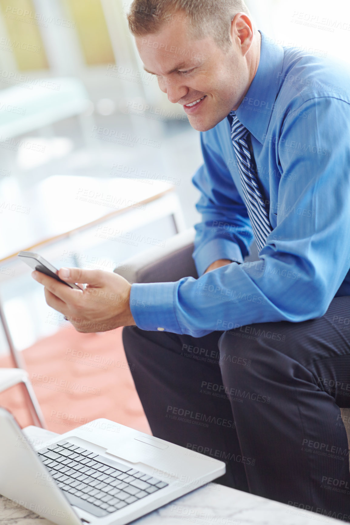 Buy stock photo A young caucasian businessman using his smartphone with his laptop before him as he works