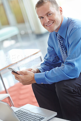 Buy stock photo A young caucasian businessman using his smartphone with his laptop before him as he works