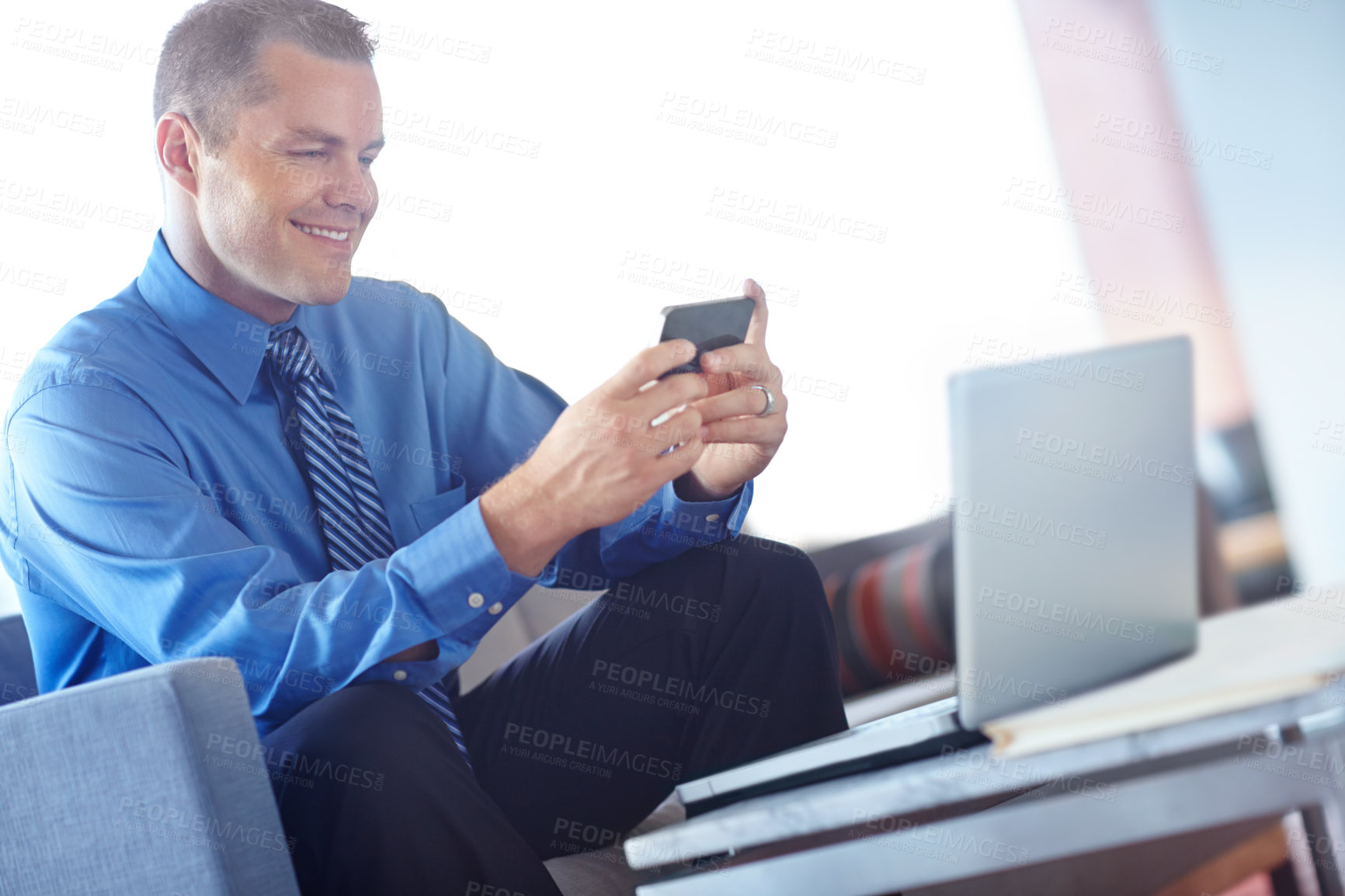 Buy stock photo A young caucasian businessman using his smartphone with his laptop before him as he works