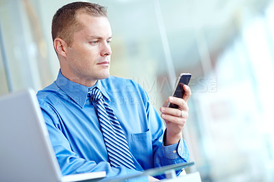 Buy stock photo A young caucasian businessman using his smartphone with his laptop before him as he works