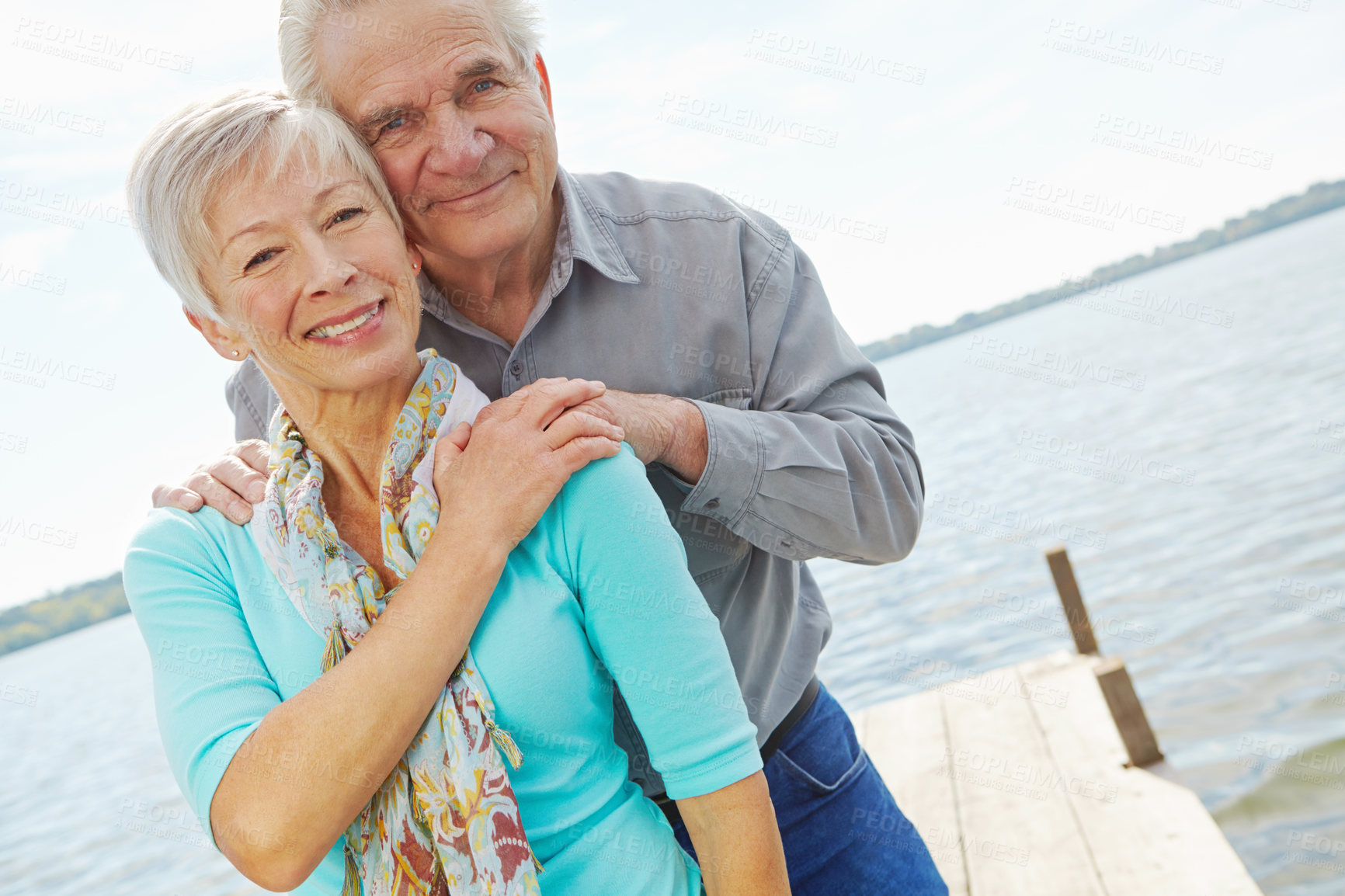 Buy stock photo A mature husband and wife looking at the camera and smiling while standing on a jetty by the lake