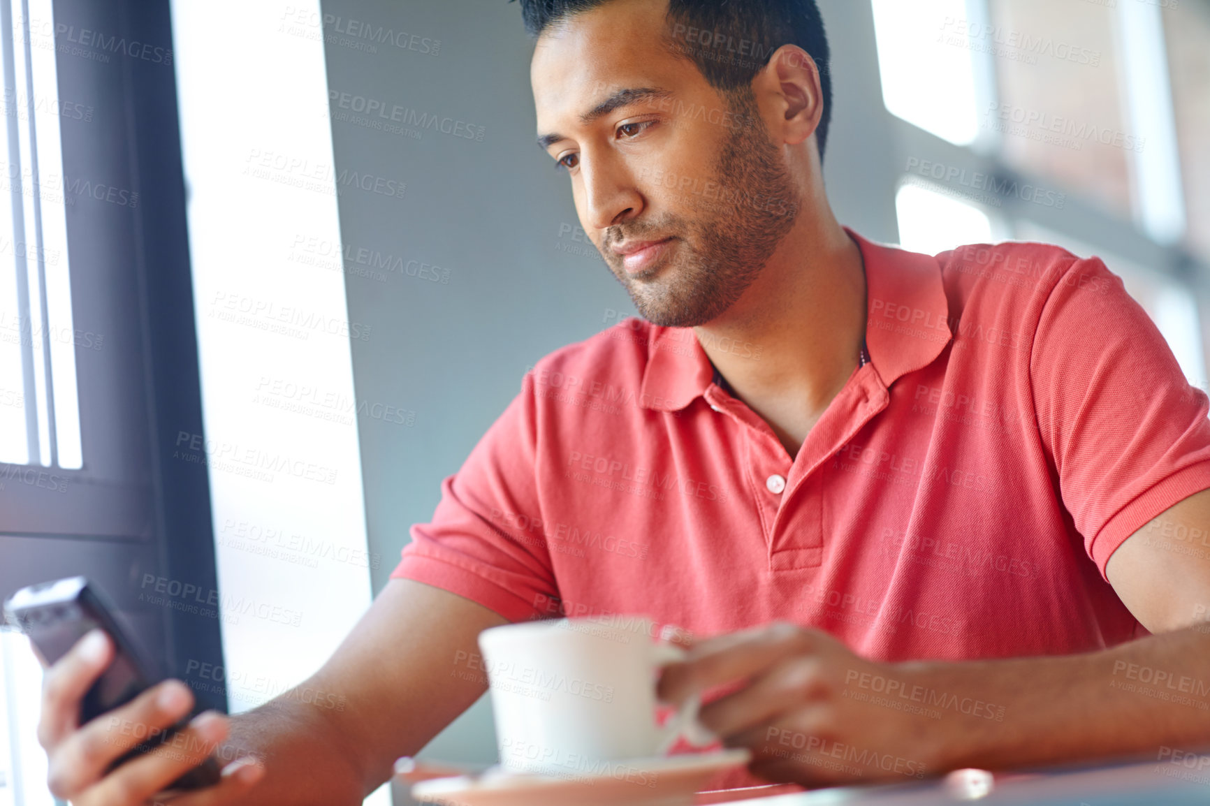 Buy stock photo A young man reading a text message while enjoying a cup of coffee