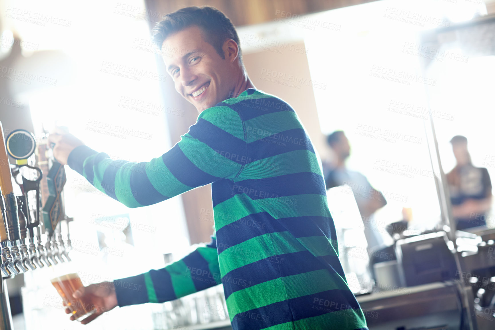 Buy stock photo A handsome young bartender pouring a pint of ale and giving you a wide smile