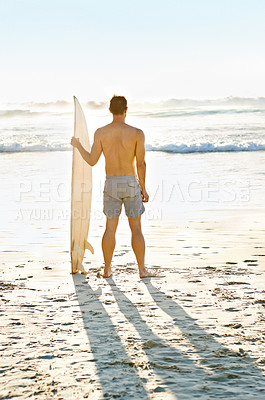 Buy stock photo Rearview shot of a muscular young man holdng his surfboard while on the beach