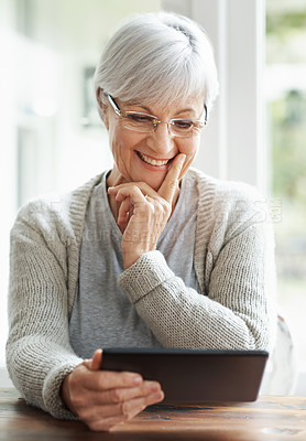 Buy stock photo A happy senior woman sitting at her kitchen table using a digital tablet