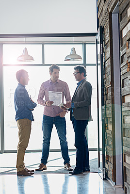 Buy stock photo Shot of male coworkers talking while standing in front of a window in an office