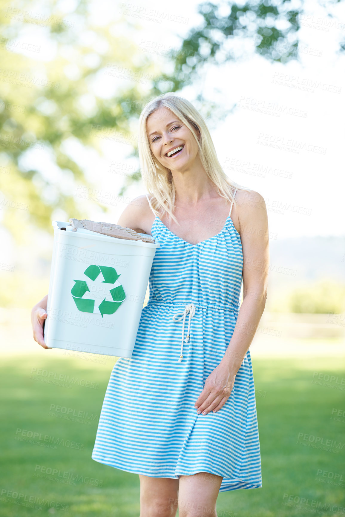 Buy stock photo A beautiful blonde woman standing in a park holding a recycling bin while smiling at the camera