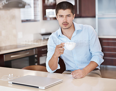 Buy stock photo A handsome male sitting in his kitchen holding a cup of coffee with a laptop in front of him