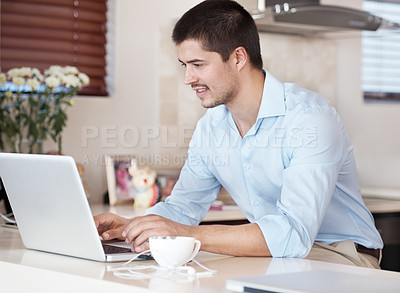 Buy stock photo A handsome male sitting in his kitchen with a cup of coffee browsing on his laptop
