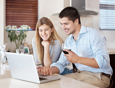 Buy stock photo A happy couple enjoying a glass of wine in their kitchen looking on a laptop together