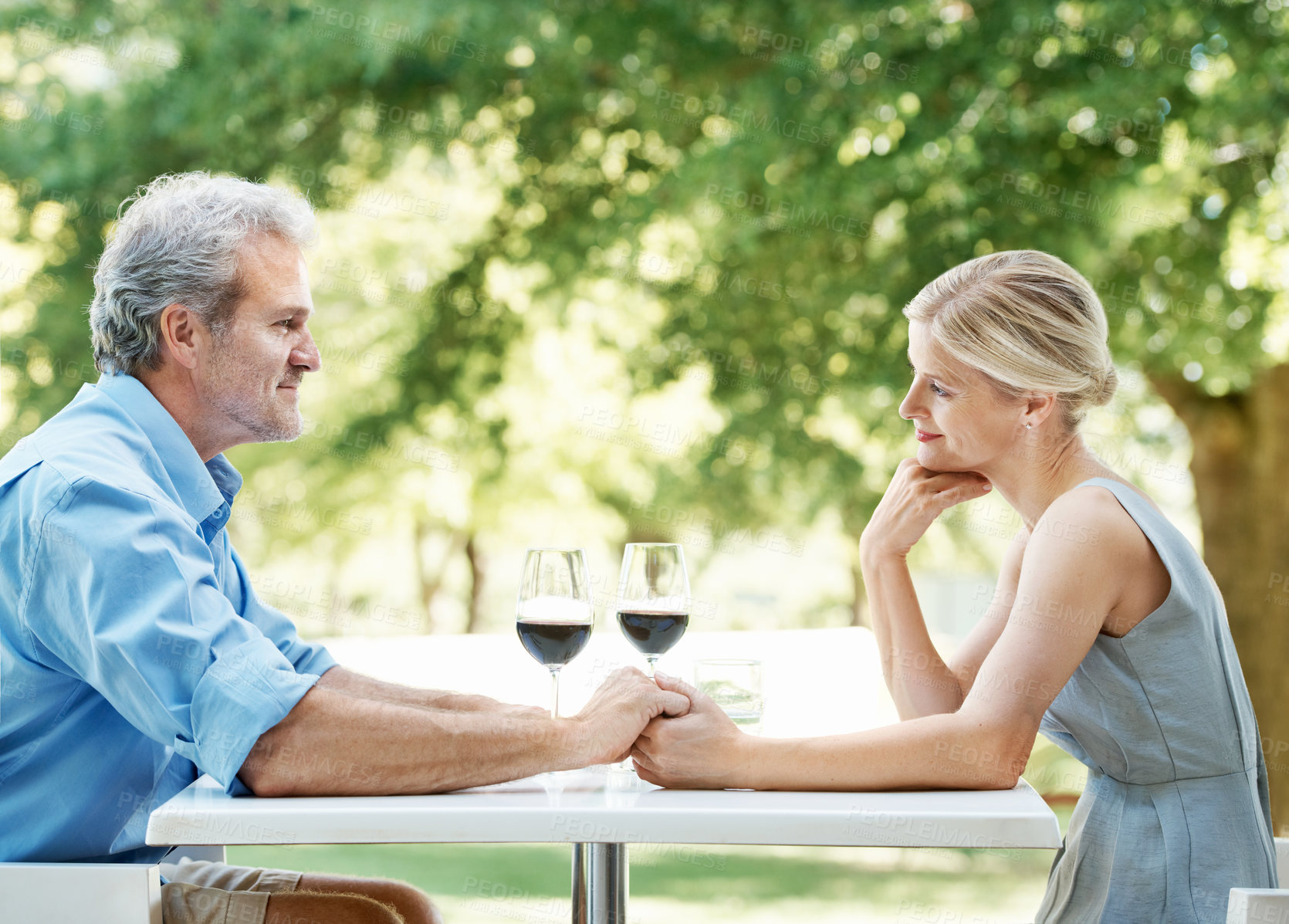 Buy stock photo Happy mature couple sitting at a table with two glasses of wine while holding hands outdoors