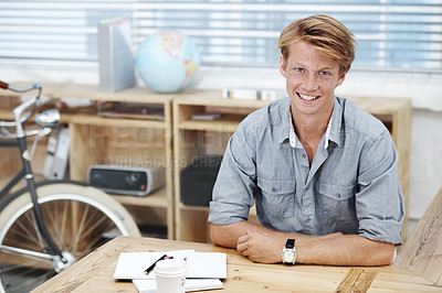 Buy stock photo Portrait of a young business man sitting in his office with wooden shelves and a bicycle. One male corporate worker seated at his desk with a cup of coffee and notebook, excited to work at his new job