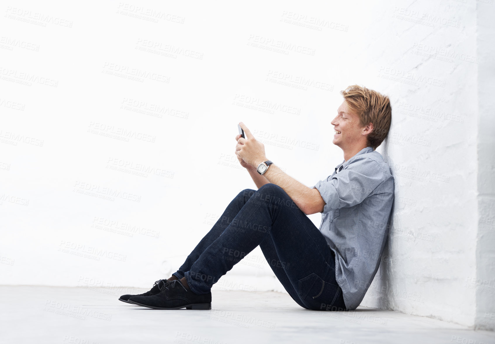 Buy stock photo Man typing on his phone smiling and sitting with copyspace. Male texting with his cellphone outside against a wall. Guy looking at social media using his mobile device connected to wifi or internet